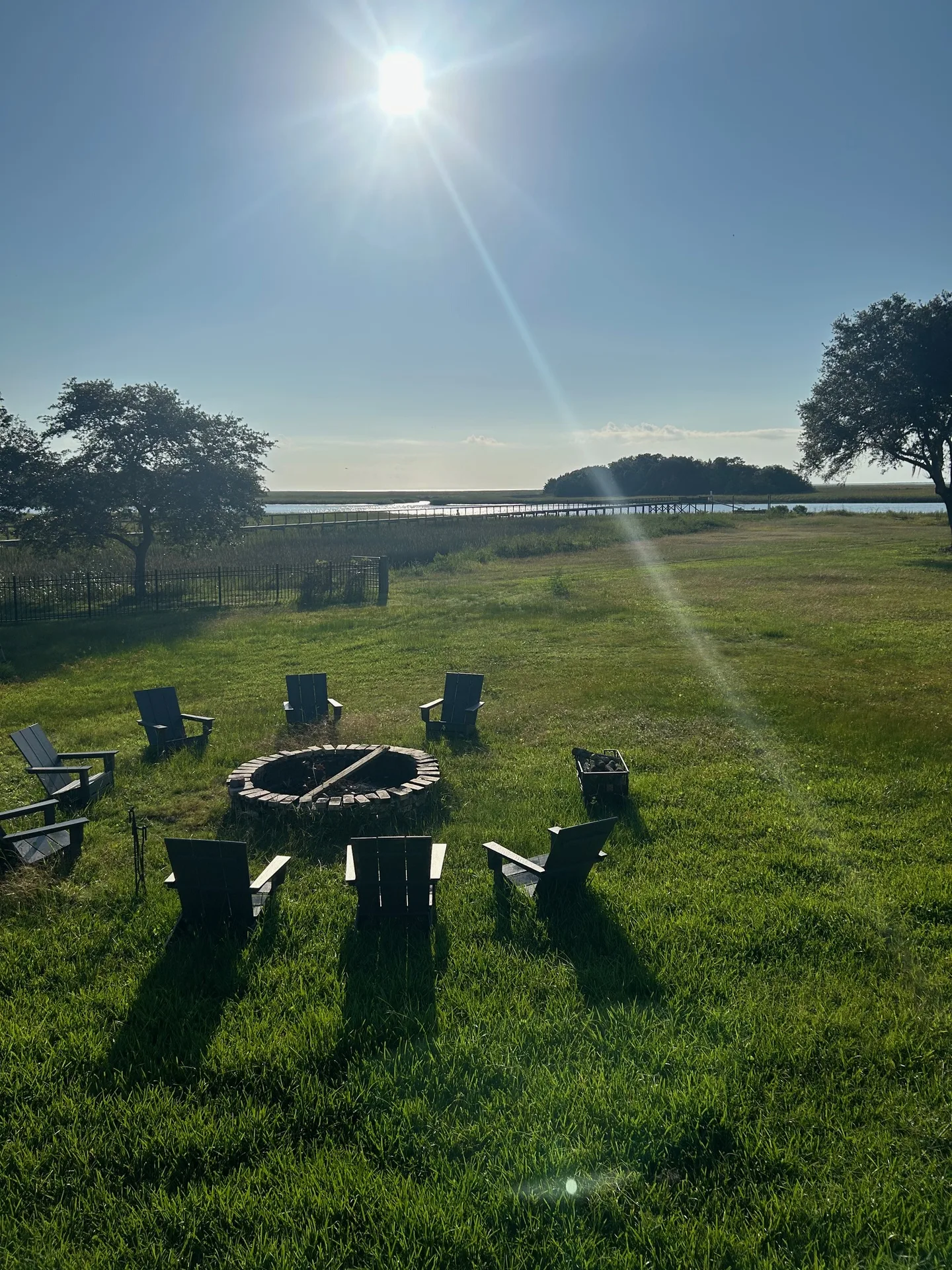 Fire pit with Adirondack chairs viewed straight-on with backlit oaks and marsh beyond
